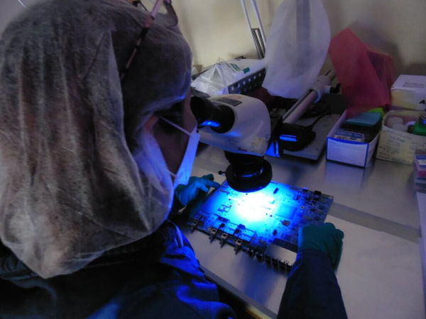 a person in protective gear uses a microscope to inspect the coating on a circuit board 